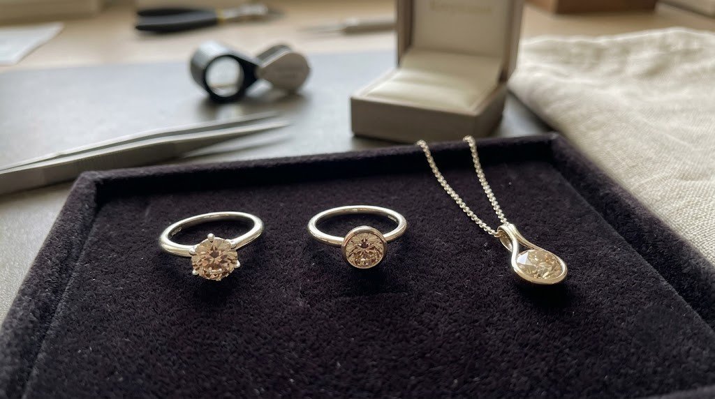 Daughter memorial diamond settings laid out on a jeweler's tray: a bezel ring, a six-prong ring and a pendant, each holding a champagne-toned stone 